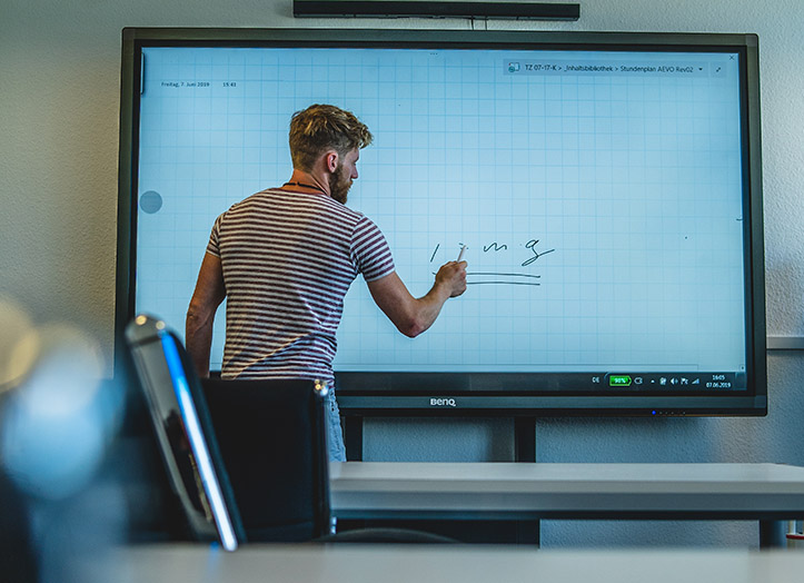 Eine Person in einem gestreiften Hemd schreibt auf einem großen interaktiven Whiteboard in einem Klassenzimmer, im Vordergrund sind Tische und Stühle zu sehen. IFF Akademie - kaufmännische Weiterbildung